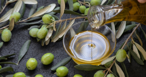 Versamento di olio extravergine di oliva in una ciotola trasparente, circondato da olive verdi fresche e ramoscelli di ulivo su sfondo scuro.