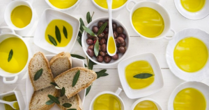 Degustazione di olio extravergine di oliva con ciotole bianche riempite di olio dorato, pane rustico, olive nere e foglie di ulivo su un tavolo bianco.