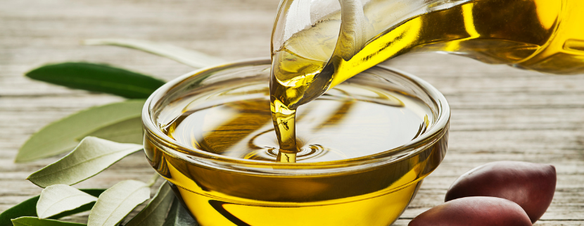 Primo piano di una caraffa di vetro che versa olio d'oliva dorato in una ciotola trasparente, circondata da foglie di ulivo e due olive mature su un tavolo di legno. L'immagine trasmette un senso di freschezza e autenticità legato alla cultura gastronomica mediterranea.