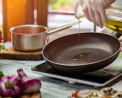 Una mano con guanto bianco versa olio extravergine di oliva in una padella antiaderente su un fornello a induzione, circondata da verdure fresche e ingredienti da cucina.