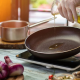 Una mano con guanto bianco versa olio extravergine di oliva in una padella antiaderente su un fornello a induzione, circondata da verdure fresche e ingredienti da cucina.