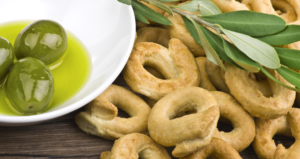 Taralli pugliesi croccanti accompagnati da olive verdi immerse in olio extravergine d’oliva, decorati con ramoscelli di ulivo su sfondo in legno rustico.