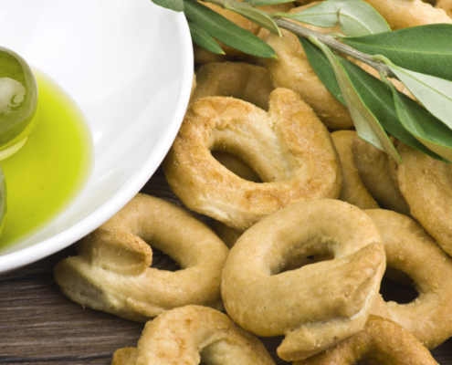 Taralli pugliesi croccanti accompagnati da olive verdi immerse in olio extravergine d’oliva, decorati con ramoscelli di ulivo su sfondo in legno rustico.