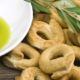 Taralli pugliesi croccanti accompagnati da olive verdi immerse in olio extravergine d’oliva, decorati con ramoscelli di ulivo su sfondo in legno rustico.