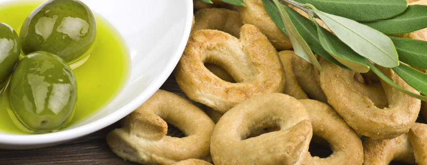 Taralli pugliesi croccanti accompagnati da olive verdi immerse in olio extravergine d’oliva, decorati con ramoscelli di ulivo su sfondo in legno rustico.