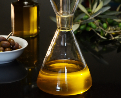Boccetta di vetro contenente olio extravergine di oliva su una superficie nera lucida, affiancata da un ciotolino di olive miste e rami di ulivo freschi.