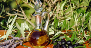 Bottiglia in vetro di olio extravergine di oliva su un tavolo all’aperto, circondata da rami d’ulivo, olive nere e fiori di lavanda, in un contesto naturale.