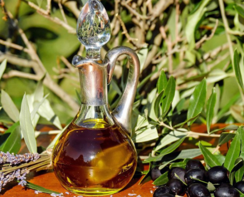 Bottiglia in vetro di olio extravergine di oliva su un tavolo all’aperto, circondata da rami d’ulivo, olive nere e fiori di lavanda, in un contesto naturale.