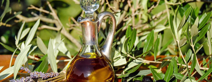 Bottiglia in vetro di olio extravergine di oliva su un tavolo all’aperto, circondata da rami d’ulivo, olive nere e fiori di lavanda, in un contesto naturale.