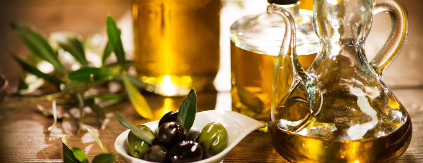 Bottiglie di olio extravergine di oliva accanto a un piattino con olive verdi e nere, su un tavolo in legno decorato con rami di ulivo, in un'atmosfera calda e rustica.