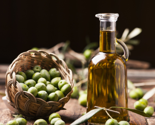 Bottiglia di olio extravergine di oliva accanto a un cestino intrecciato colmo di olive verdi, su un tavolo rustico in legno, con rami di ulivo sparsi, simbolo della tradizione e genuinità dei prodotti tipici mediterranei.