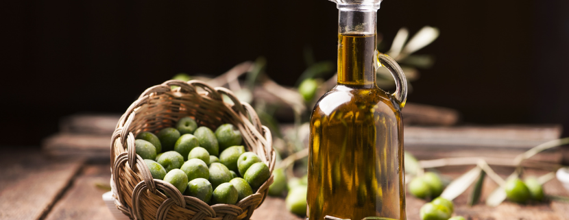 Bottiglia di olio extravergine di oliva accanto a un cestino intrecciato colmo di olive verdi, su un tavolo rustico in legno, con rami di ulivo sparsi, simbolo della tradizione e genuinità dei prodotti tipici mediterranei.