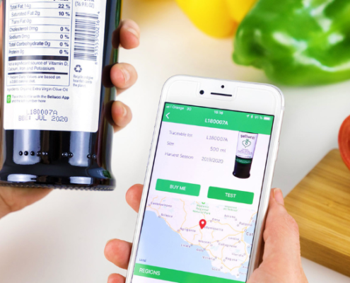 Persona che utilizza uno smartphone per tracciare l’origine di una bottiglia di olio extravergine tramite app blockchain, visualizzando i dati di produzione e la posizione geografica sulla mappa. Sullo sfondo verdure fresche su un piano da cucina.