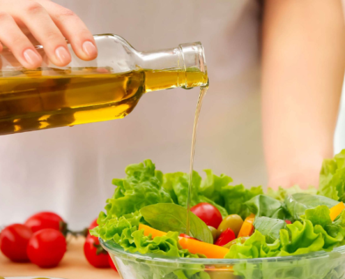 Persona che versa olio extravergine di oliva da una bottiglia su una ciotola di insalata fresca con lattuga, pomodorini e verdure colorate.