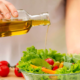 Persona che versa olio extravergine di oliva da una bottiglia su una ciotola di insalata fresca con lattuga, pomodorini e verdure colorate.