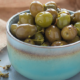 Ciotola in ceramica azzurra colma di olive verdi condite, appoggiata su un panno di juta, pronta per essere servita come antipasto o snack mediterraneo.