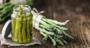 Vasetto di asparagi selvatici sott’olio su un tavolo in legno rustico, con mazzetti di asparagi freschi legati con spago naturale sullo sfondo, a rappresentare un prodotto artigianale e genuino della tradizione contadina.