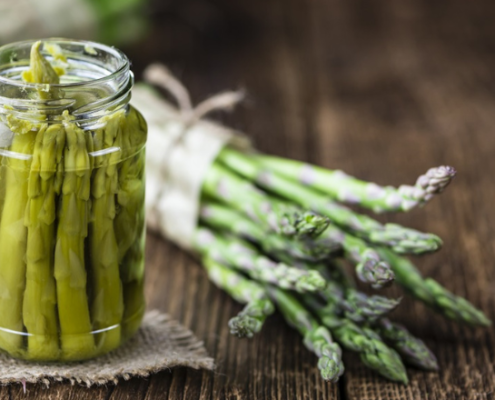 Vasetto di asparagi selvatici sott’olio su un tavolo in legno rustico, con mazzetti di asparagi freschi legati con spago naturale sullo sfondo, a rappresentare un prodotto artigianale e genuino della tradizione contadina.