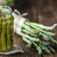 Vasetto di asparagi selvatici sott’olio su un tavolo in legno rustico, con mazzetti di asparagi freschi legati con spago naturale sullo sfondo, a rappresentare un prodotto artigianale e genuino della tradizione contadina.