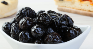 Ciotola bianca con olive nere al forno, dalla superficie rugosa e lucida, pronte per essere gustate come antipasto o condimento tipico della cucina mediterranea, su sfondo chiaro con pane o focaccia sullo sfondo sfocato.