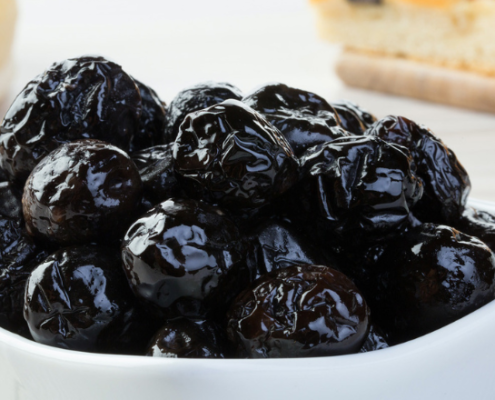 Ciotola bianca con olive nere al forno, dalla superficie rugosa e lucida, pronte per essere gustate come antipasto o condimento tipico della cucina mediterranea, su sfondo chiaro con pane o focaccia sullo sfondo sfocato.