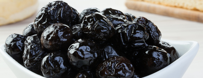 Ciotola bianca con olive nere al forno, dalla superficie rugosa e lucida, pronte per essere gustate come antipasto o condimento tipico della cucina mediterranea, su sfondo chiaro con pane o focaccia sullo sfondo sfocato.