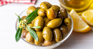Ciotola di olive verdi schiacciate condite con olio extravergine di oliva e decorate con una foglia di ulivo, accompagnata da spicchi di limone su un tavolo in legno chiaro.
