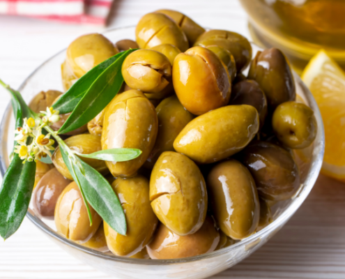 Ciotola di olive verdi schiacciate condite con olio extravergine di oliva e decorate con una foglia di ulivo, accompagnata da spicchi di limone su un tavolo in legno chiaro.