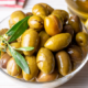Ciotola di olive verdi schiacciate condite con olio extravergine di oliva e decorate con una foglia di ulivo, accompagnata da spicchi di limone su un tavolo in legno chiaro.