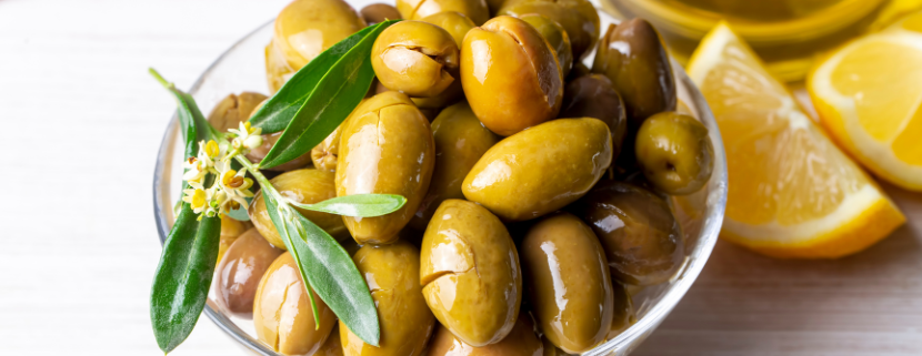 Ciotola di olive verdi schiacciate condite con olio extravergine di oliva e decorate con una foglia di ulivo, accompagnata da spicchi di limone su un tavolo in legno chiaro.