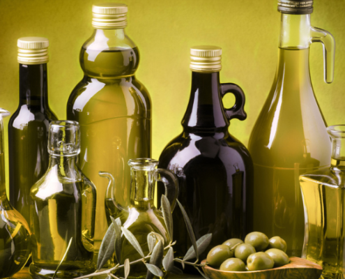 Diverse bottiglie e oliere riempite con vari tipi di olio d’oliva, disposte su un tavolo insieme a olive fresche e rametti di ulivo, su sfondo verde illuminato.