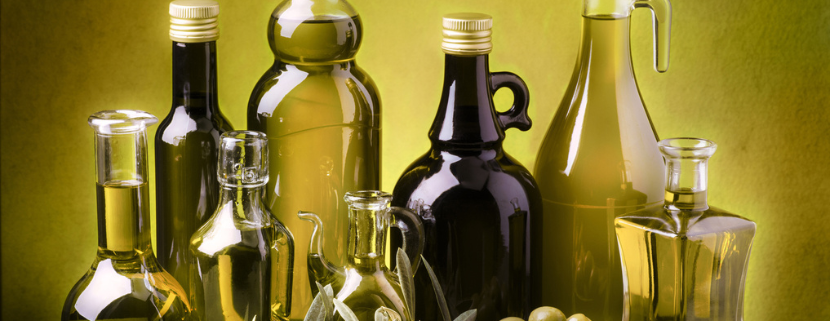 Diverse bottiglie e oliere riempite con vari tipi di olio d’oliva, disposte su un tavolo insieme a olive fresche e rametti di ulivo, su sfondo verde illuminato.