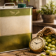 Lattina in metallo per la conservazione dell’olio extravergine di oliva, con olive verdi, ramoscelli d’ulivo, una ciotola di olio e un orologio antico su tavolo in legno.