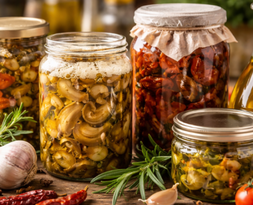 Barattoli di conserve sott’olio con verdure, funghi e pomodori secchi su un tavolo rustico, accanto a una bottiglia di olio extravergine di oliva, in formato orizzontale senza testo.