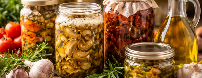 Barattoli di conserve sott’olio con verdure, funghi e pomodori secchi su un tavolo rustico, accanto a una bottiglia di olio extravergine di oliva, in formato orizzontale senza testo.
