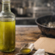 Bottiglia di olio d'oliva aperta su un tavolo in legno accanto a un cucchiaio con olio e a un cartoncino con data di scadenza, mentre sullo sfondo una padella con olio caldo produce fumo in cucina.