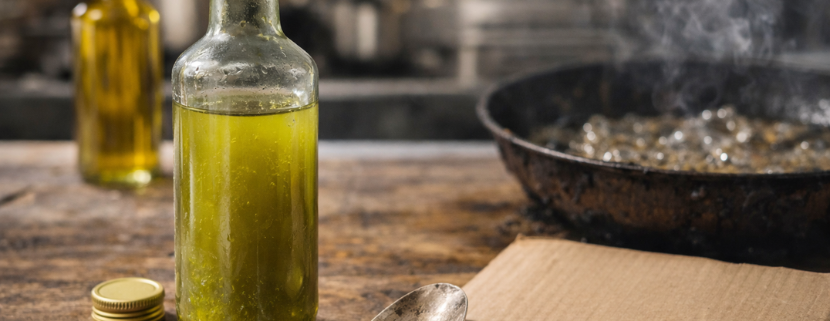 Bottiglia di olio d'oliva aperta su un tavolo in legno accanto a un cucchiaio con olio e a un cartoncino con data di scadenza, mentre sullo sfondo una padella con olio caldo produce fumo in cucina.